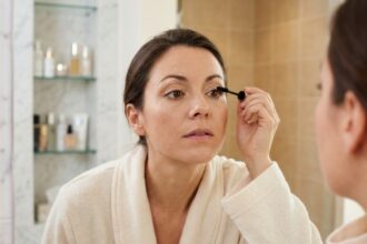 Femme portant des lentilles de contact appliquant du mascara devant un miroir de salle de bain, routine maquillage de fêtes sans irritation
